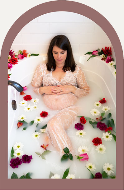 Pregnant woman wearing long dress laying in bathtub filled with milk and flowers.